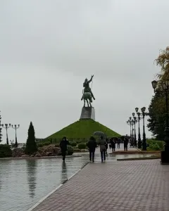 The Salavat Yulaev Monument stands on a grassy mound above a wet walkway, with visitors holding umbrellas on a rainy day.