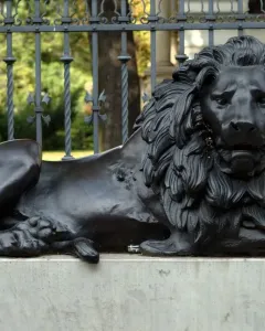 Guarding Lion sculpture on a concrete pedestal in front of an ornate fence and brick pillar.