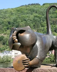 Heidelberg Bridge Monkey sculpture crouching on a stone ledge, holding a round disk with the river and wooded hill behind it.