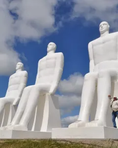 The Man by the Sea sculpture with four massive seated white figures against a blue sky, with a person beside the front statue.