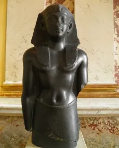 Upper body of a Ptolemaic king statue in dark stone, wearing an Egyptian headcloth and kilt with broken arms.