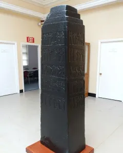 Black Obelisk with stepped top and carved relief bands on a wooden base in a museum interior.