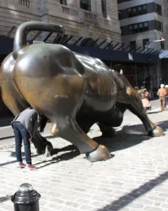 Charging Bull bronze sculpture seen from the rear on a cobblestone street with pedestrians nearby.