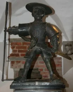 Old Thomas statue in armor with a wide-brimmed hat, mustache, sword, and long pole-like weapon.