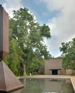 Barnett Newman’s Broken Obelisk sculpture rising from a reflecting pool with trees and a light-toned building behind it.