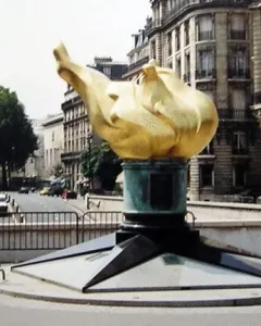 The Flame of Liberty sculpture with a golden flame on a dark stepped base in an urban street setting.