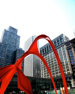 Flamingo, a large red abstract steel sculpture, stands in a city plaza with tall buildings in the background and a person near its base.