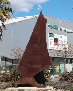Dorna, a rust-colored abstract metal sculpture with a pointed top and splayed base, set on a stone wall among shrubs.