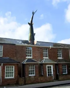 The Headington Shark sculpture plunging into the roof of a red brick building under a blue sky.