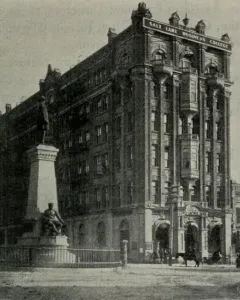The Brigham Young Monument with a bronze figure atop a tall stone pedestal and relief sculptures at the base, with a white building and bare trees behind it.