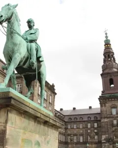 Christian IX equestrian statue on a tall stone pedestal in an open square, with ornate buildings and a slender tower behind it.