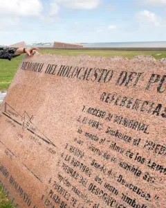 A woman stands beside the Holocaust Memorial plaque, a large angled pink granite monument with engraved text, near a grassy waterfront.