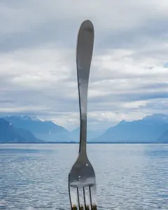 The Fork of Vevey sculpture stands upright in calm water with its reflection visible.