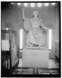 A large seated marble statue of a male figure with one arm raised and the other holding a bound object, displayed in a room with arched windows.