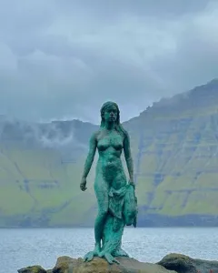 Kópakonan bronze statue standing on dark rocks by the water with misty mountains behind it.