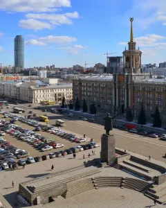 Lenin Monument stands on a tall pedestal in a city square with steps, people, cars, and surrounding buildings.