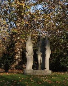 Three Standing Figures sculpture standing on a low base on a grassy lawn beside a tree.