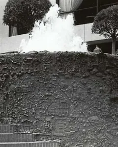 San Francisco Fountain with water cascading from its top edge in front of a modern building and two small trees.