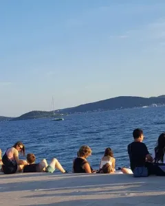 People sitting on the Sea Organ stone steps by the blue sea in Zadar, looking toward distant islands and a sailboat.