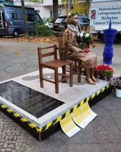 Peace Statue showing a bronze girl seated barefoot on a stool beside an empty wooden chair, holding a bouquet of flowers.