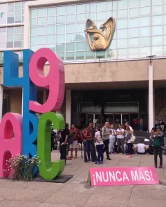 Antimonumento 49 ABC stands on a paved plaza in front of a modern building, with a small bouquet of white flowers and a pink sign reading \