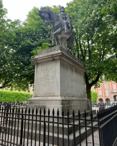 Equestrian statue of Louis XIII on a tall stone pedestal behind a black fence, with trees and a brick building in the background.