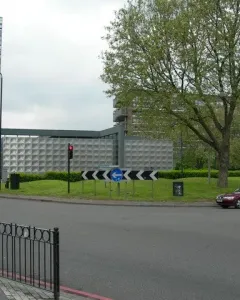 Michael Faraday Memorial on a grassy roundabout island beside roads and traffic lights, with buildings and trees in the background.