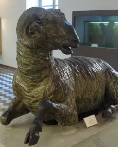 Bronze ram sculpture with curled horns resting on a stone pedestal in a museum gallery.