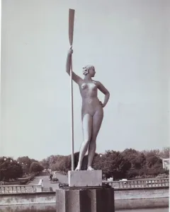 Girl with an Oar sculpture standing on a tiered pedestal in a park fountain, holding a tall oar upright.