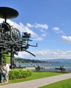 Heureka sculpture standing on a grassy lawn beside Lake Zurich, with mountains in the distance.