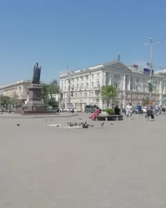 The Monument to Dimitry of Rostov stands on a tiered pedestal in a city square, with pedestrians, pigeons, and buildings in the background.