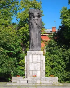 Karl Marx monument with a dark stone figure on a tiered base, framed by green trees and a brick building.