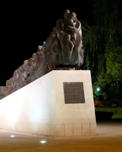 The Train of Pain monument at night, showing a long dark sculpture of clustered human figures on a light-colored base.