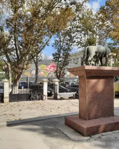 Capitoline Wolf sculpture with Romulus and Remus on a tall red-stone pedestal in an urban square.