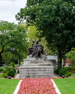 Wars of America bronze sculpture on a stone base in a green park with trees and a flower-lined path.