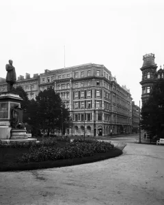 Johan Ludvig Runeberg statue on a large stone pedestal along a park pathway, with historic buildings and trees behind it.