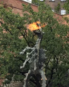 Wawel Dragon sculpture spewing a bright flame from its open mouth beside trees and a brick building