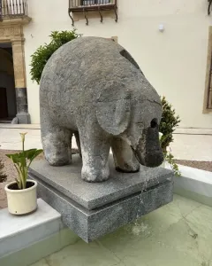 Stone elephant fountain standing on a granite base with water flowing from its trunk into a shallow basin.