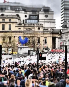 The blue Hahn/Cock sculpture stands on a pedestal in Trafalgar Square with a large crowd nearby.