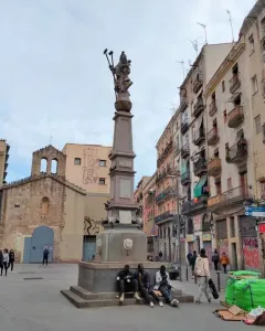 Font de Santa Eulàlia, a tall stone fountain monument in a city square with people seated around its base.