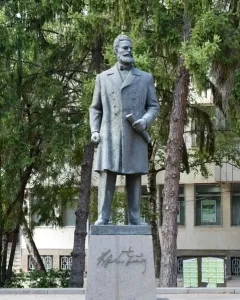 Monument to Hristo Botev: a bronze-colored statue of a bearded man holding a rolled document on a tall stone pedestal among trees.