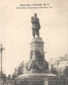 Monument of Alexander III on an ornate pedestal with a decorative iron fence in front of a classical building.