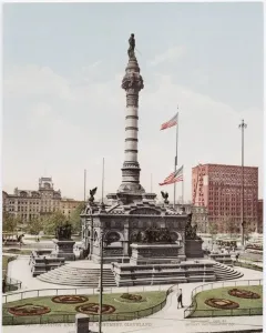 Soldiers\' and Sailors\' Monument rising from a stepped base in a landscaped park, with American flags nearby and city buildings in the background.