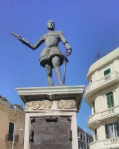 Bronze statue of John of Austria in historical armor on a tall stone pedestal, with one arm extended and a sword in the other hand.