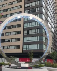 The Eye of Mexico sculpture in an urban plaza beside a tall gridded building, framed by trees and city streets.