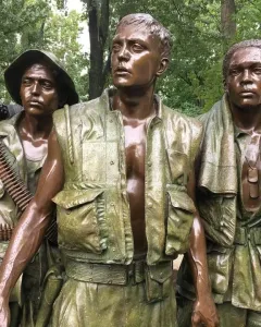 The Three Soldiers bronze statue with three wet-looking soldiers standing outdoors among trees.