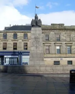 Paisley War Memorial stone cenotaph in a town square with carved details and dark figures on top.