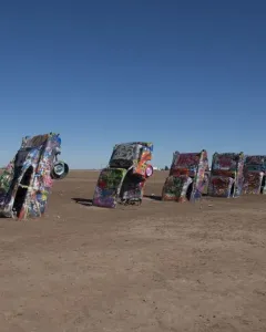Cadillac Ranch cars covered in graffiti stand buried nose-first in a flat dirt field under a clear blue sky.