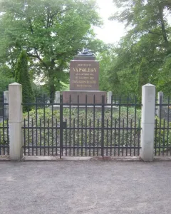 Napoleonstein stone monument behind a dark metal fence and stone pillars, surrounded by trees and greenery.
