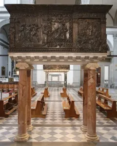 The Passion Pulpit in a church interior, with dark bronze relief carvings on red marble columns above a checkered floor and wooden pews.